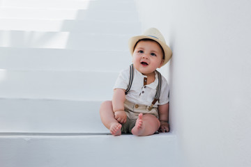 Beautiful baby sitting on white stairs in Santorini, Greece, wearing a hat and a vintage outfit
