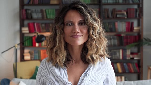 Beautiful curly woman in thirties of fair complexion in white casual shirt looks softly into camera and smiles gently in stylish spacious room with sophisticated decor in front of a large bookcase