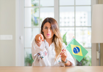 Young woman at home holding flag of Brazil pointing with finger to the camera and to you, hand...