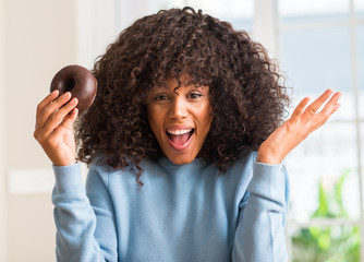 Young african american woman holding chocolate donut at home very happy and excited, winner expression celebrating victory screaming with big smile and raised hands