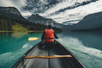 Young Man Canoeing on Emerald Lake in the rocky mountains canada with canoe and life vest with mountains in the background blue water. - obrazy, fototapety, plakaty