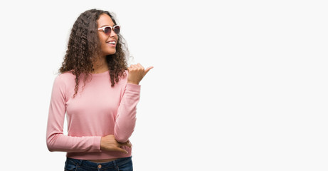 Beautiful young hispanic woman wearing sunglasses smiling with happy face looking and pointing to...