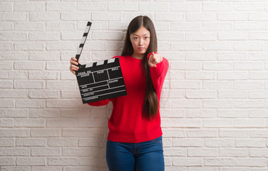Young Chinese woman over white brick wall holding clapboard pointing with finger to the camera and...