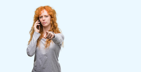 Young redhead woman talking on the phone pointing with finger to the camera and to you, hand sign, positive and confident gesture from the front