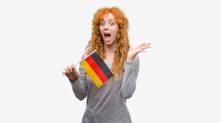 Young redhead woman holding flag of Germany very happy and excited, winner expression celebrating victory screaming with big smile and raised hands