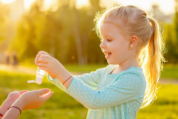 cute little blonde girl daughter in a blue dress applying antiseptic antibacterial gel in summer day in field greens grass background. protection from germs at picnic and travel trip