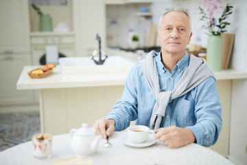 Mature man in casualwear sitting by table in the kitchen and having tea in the morning