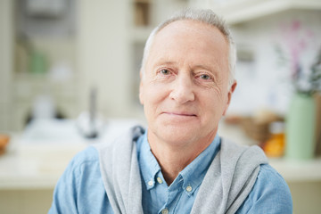 Mature grey-haired man in denim shirt looking at you while sitting in the kitchen