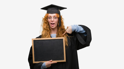Young redhead student woman wearing graduated uniform holding blackboard with surprise face pointing finger to himself