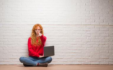 Young redhead woman sitting over brick wall using computer laptop pointing with finger to the...
