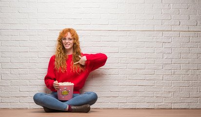 Young redhead woman sitting over brick wall eating popcorn with surprise face pointing finger to himself