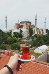 Naklejka premium Traditional Turkish tea in a glass on a plate in the hands of a man with a mosque Ayasofya background. 