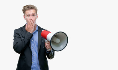 Young handsome blond man holding a megaphone cover mouth with hand shocked with shame for mistake, expression of fear, scared in silence, secret concept