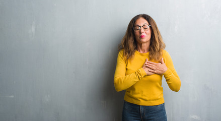 Middle age hispanic woman over grey wall wearing glasses smiling with hands on chest with closed eyes and grateful gesture on face. Health concept.