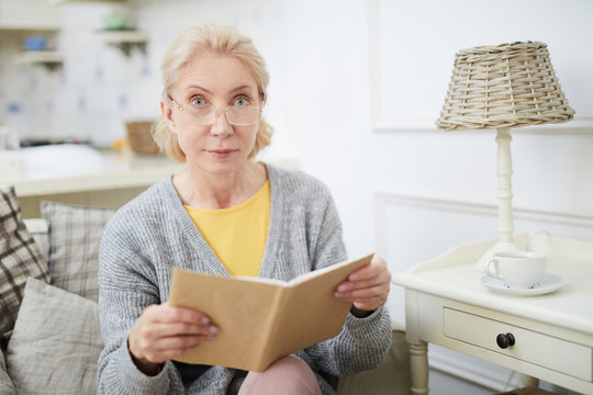 Aged serious woman in eyeglasses looking at camera while reading book at leisure at home