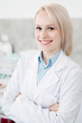 Happy young doctor in whitecoat crossing her arms on ches and looking at you with toothy smile