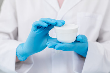 Gloved hands of modern cosmetician holding white plastic jar with facial cream