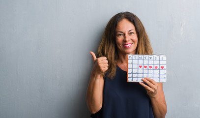 Middle age hispanic woman standing over grey grunge wall holding period calendar pointing and...