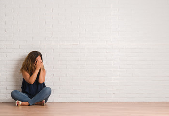 Middle age hispanic woman sitting on the floor over white brick wall with sad expression covering face with hands while crying. Depression concept.