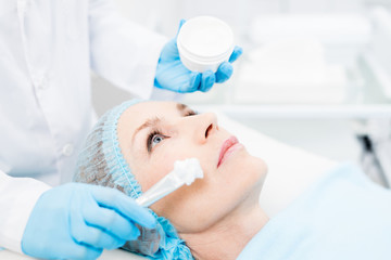 Cosmetician in whitecoat and gloves applying mask or cream on face of mature female after massage