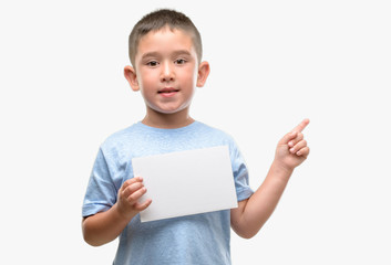 Dark haired little child holding a blank card very happy pointing with hand and finger to the side