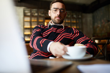 Serious man in pullover and eyeglasses sitting by table in cafe and having tea or coffee