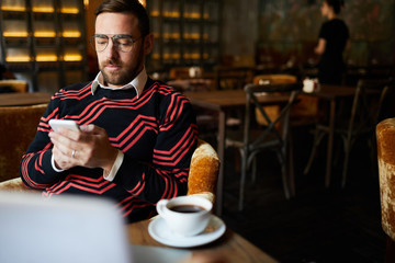 Young serious businessman in casualwear messaging in smartphone while sitting in cafe