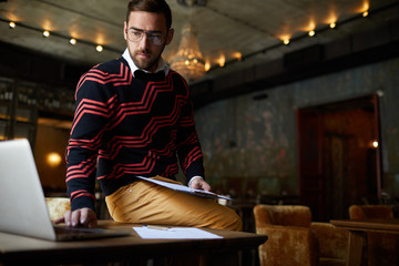 Serious businessman in casualwear sitting on table in front of laptop and networking in cafe