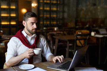 Serious businessman with drink sitting by table in front of laptop and searching for necessary data