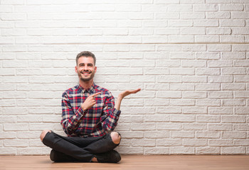 Young adult man sitting over white brick wall very happy pointing with hand and finger