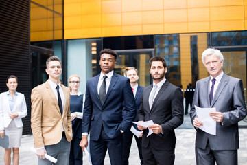 Group of confident intercultural businessmen in suits and their female colleagues standing not far...