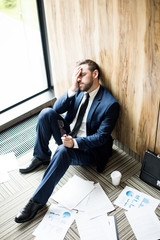 Tired or stressed businessman in suit sitting on the floor with papers around him and touching his...