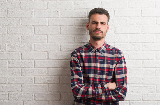 Young adult man standing over white brick wall skeptic and nervous, disapproving expression on face with crossed arms. Negative person.