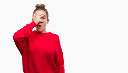 Young blonde woman wearing bun and red sweater peeking in shock covering face and eyes with hand,...