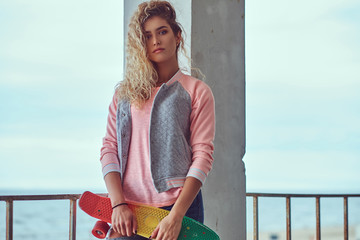 Charming young girl with blonde hair dressed in a pink jacket holds a skateboard posing near a...