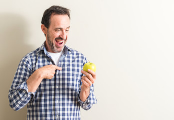 Senior man holding a green apple with surprise face pointing finger to himself