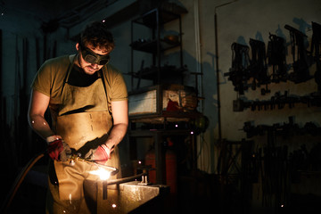 Young blacksmith in protective eyeglasses and apron standing by anvil and welding one of iron workpieces