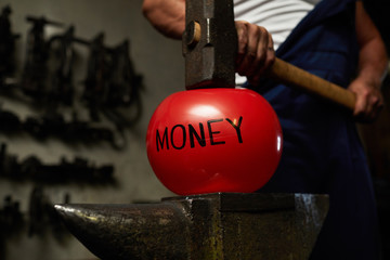 Red ball symbolizing money concept is situated on anvil with big heavy hammer above