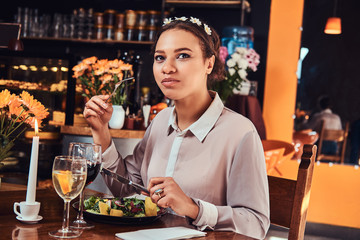 Portrait of a beautiful black-skinned woman wearing a blouse and flower headband, enjoying dinner while eating in a restaurant.
