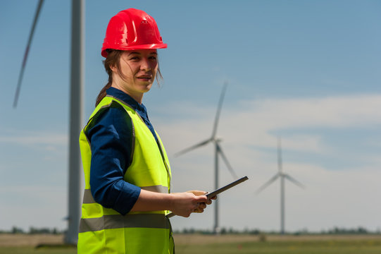 Serious successful young female engineer in a green vest and red hard hat designing a plan against a background of windmills