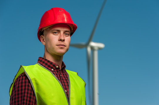 Portrait of a serious ambitious engineer wearing red hard hat against the backdrop of a windmill and a blue sky.