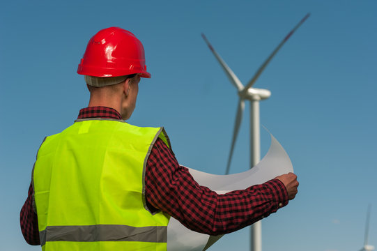 Rear view of unknown male engineer in a green waistcoat with red helmet with engineering plans against the background of windmills and the blue sky