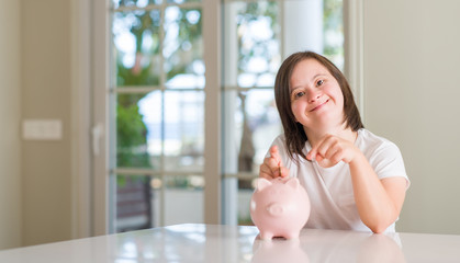 Down syndrome woman at home holding piggy bank very happy pointing with hand and finger