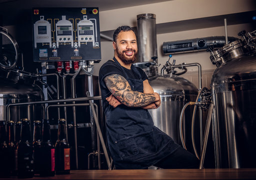 Portrait of a stylish bearded tattooed dark skinned male with crossed arms standing behind the counter in the brewery.