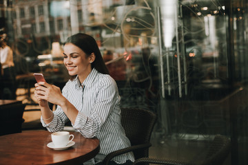 Pretty brunette is sitting at table and holding phone in hands and looking at it. She is smiling. There is a white cup on the table. Girl is in restaurant.