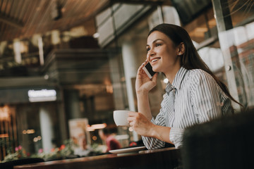 Cheerful and attractive woman is sitting at table in restaurant. She is talking on the phone. Girl is looking forward and smiling. She is happy. Young woman has a white cup in hands.