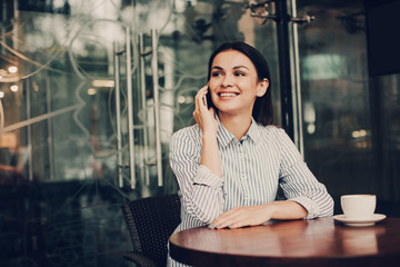 Brunette is sitting at table and looking to the right. She is holding phone close to ear and smiling. Girl is talking on the phone. There is a cup of coffee at table. Girl looks happy.