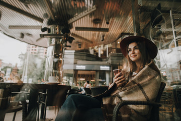 Nice view of girl sitting in chair. She has blanket on her shoulders. Woman is holding cup of coffee and enjoying the moment. Girl is looking forward. She is istting in restaurant.