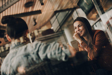 Cheerful girl in brown jacket is sitting at table with her friend and smiling. Girls are sitting in front of each other. They are talking and having conversation. Women are in restaurant.
