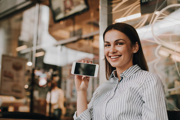 Positive and pretty girl is sitting at table and looking on camera. She is smiling. Woman is showing phone's screen on camera. Girl is sitting near glass window.